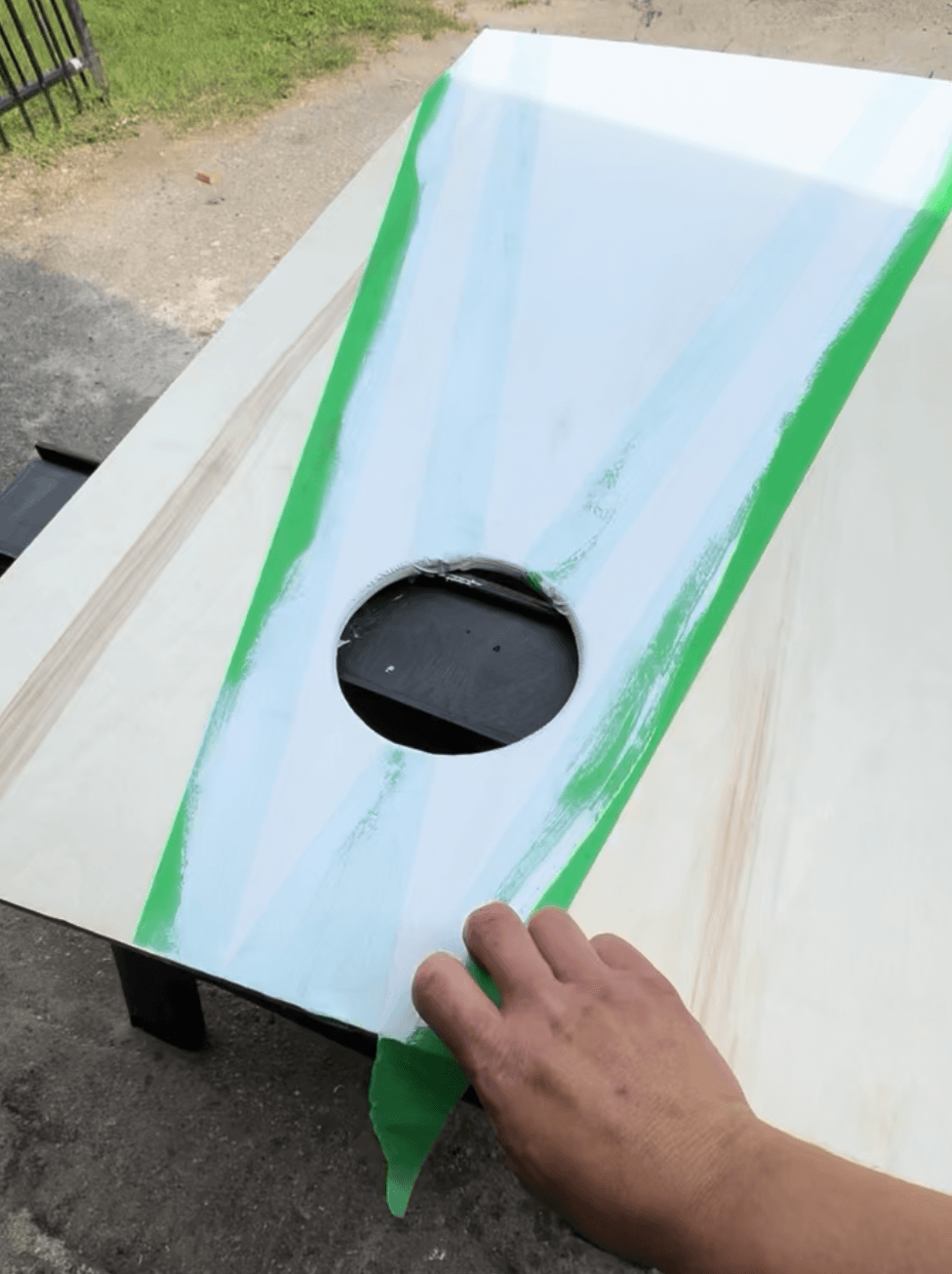 Hand peeling green tape off a freshly painted cornhole board revealing a clean painted edge around the hole