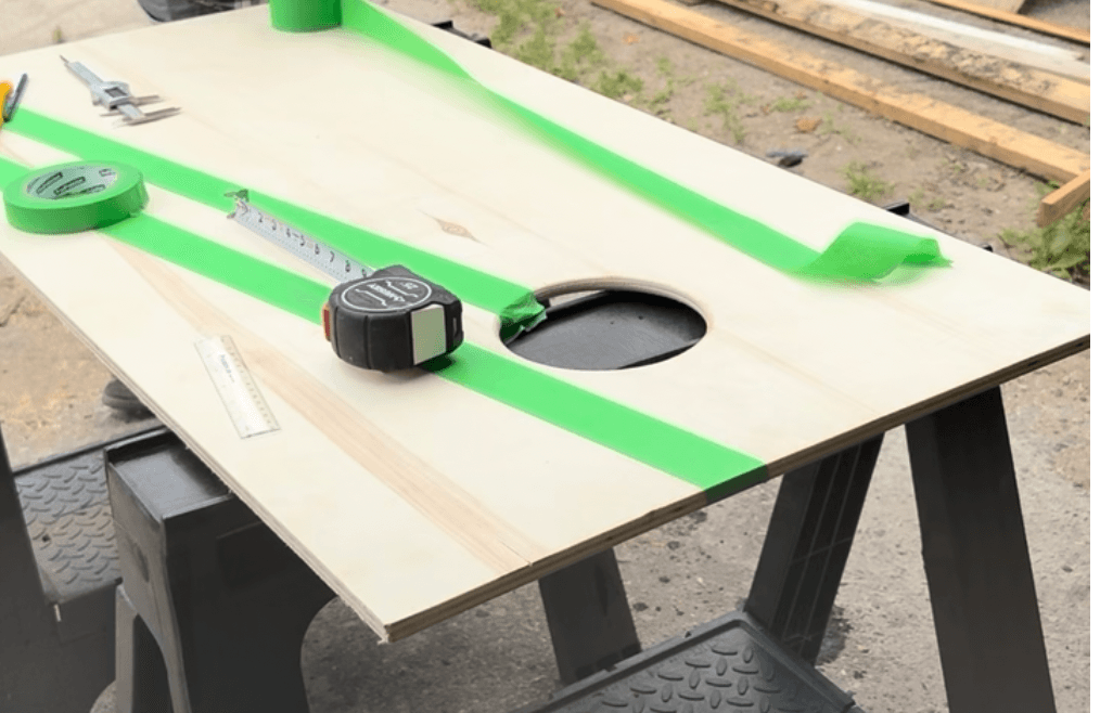 Cornhole board masked with green painter's tape before painting, hole visible in center