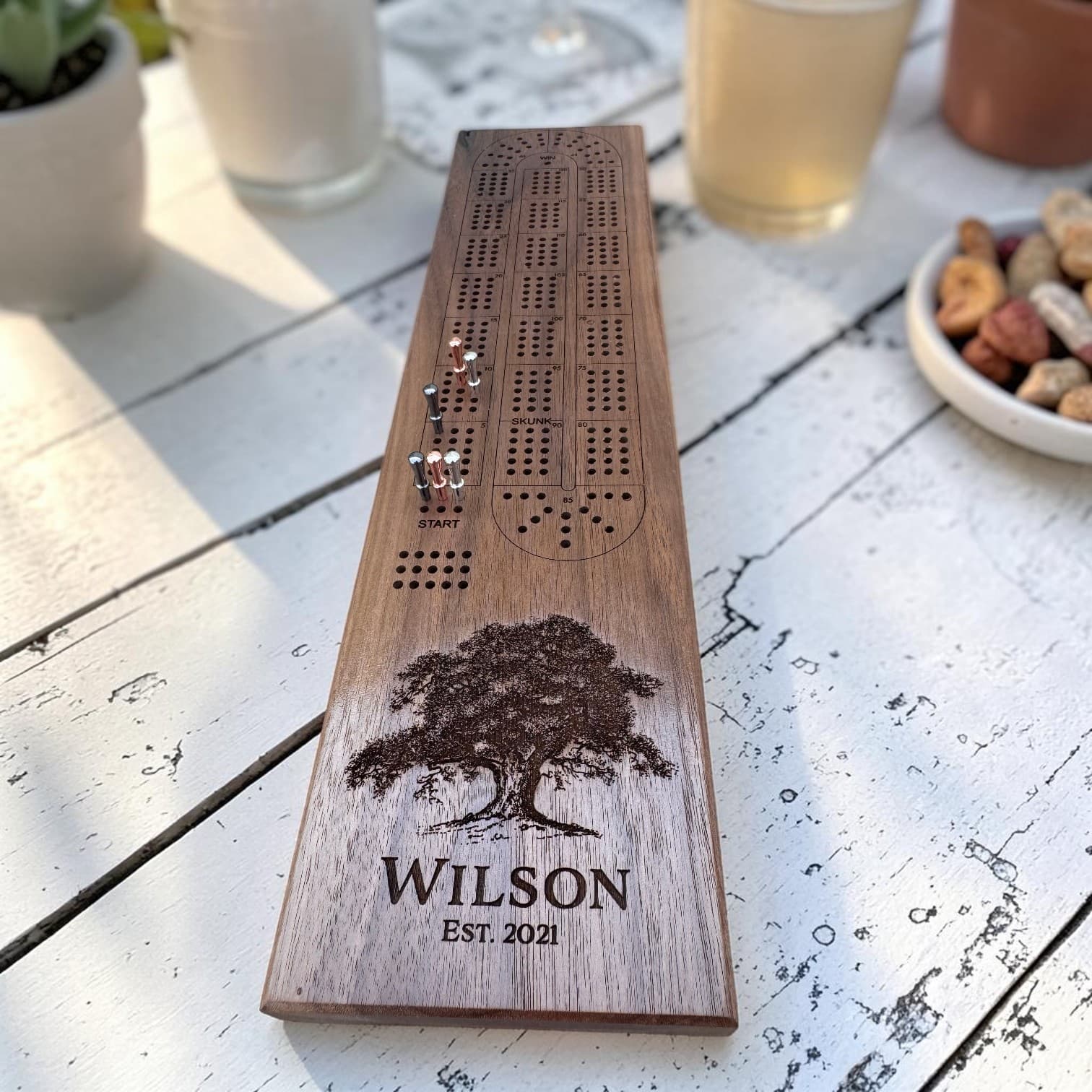 Personalized Black Walnut cribbage board with pegs in play on a patio table, engraved with a family name, est. year, and hand-drawn tree