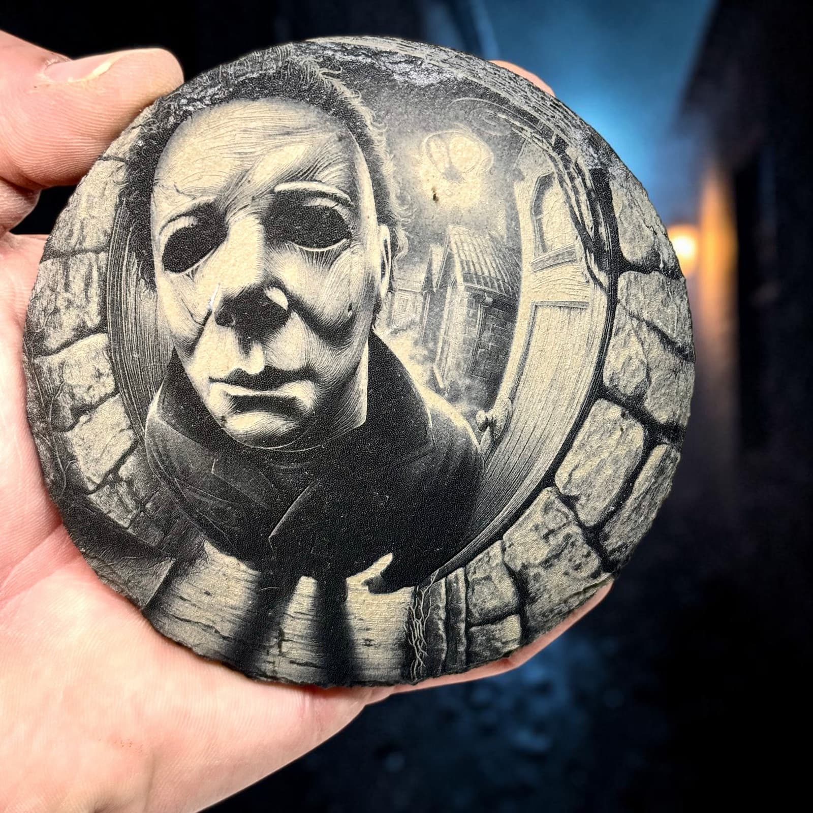 Round slate coaster engraved with Michael Myers seen through a fisheye peephole, held in a hand for scale
