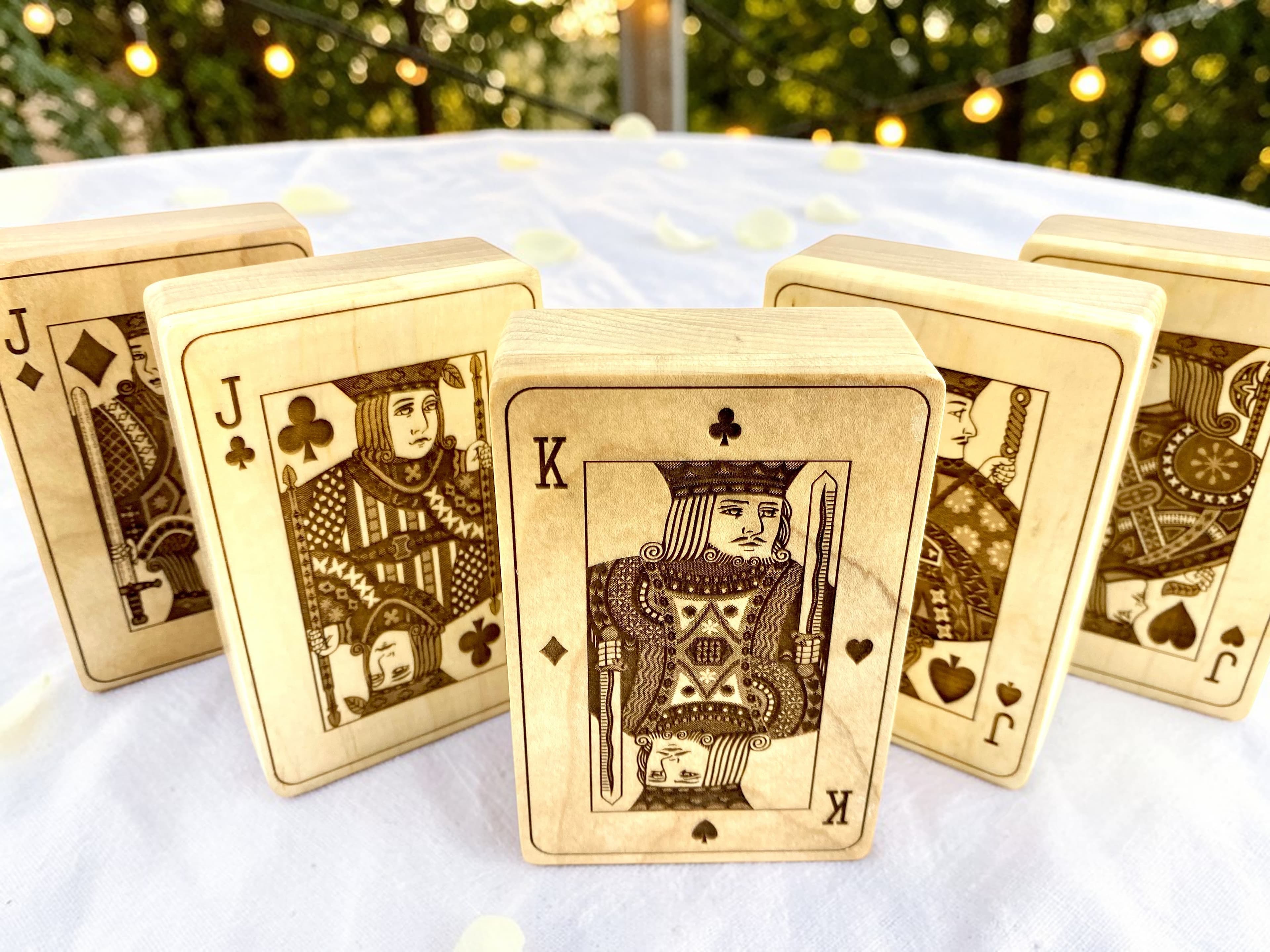 Five custom engraved maple playing card boxes standing upright at an outdoor wedding reception with string lights and greenery in the background
