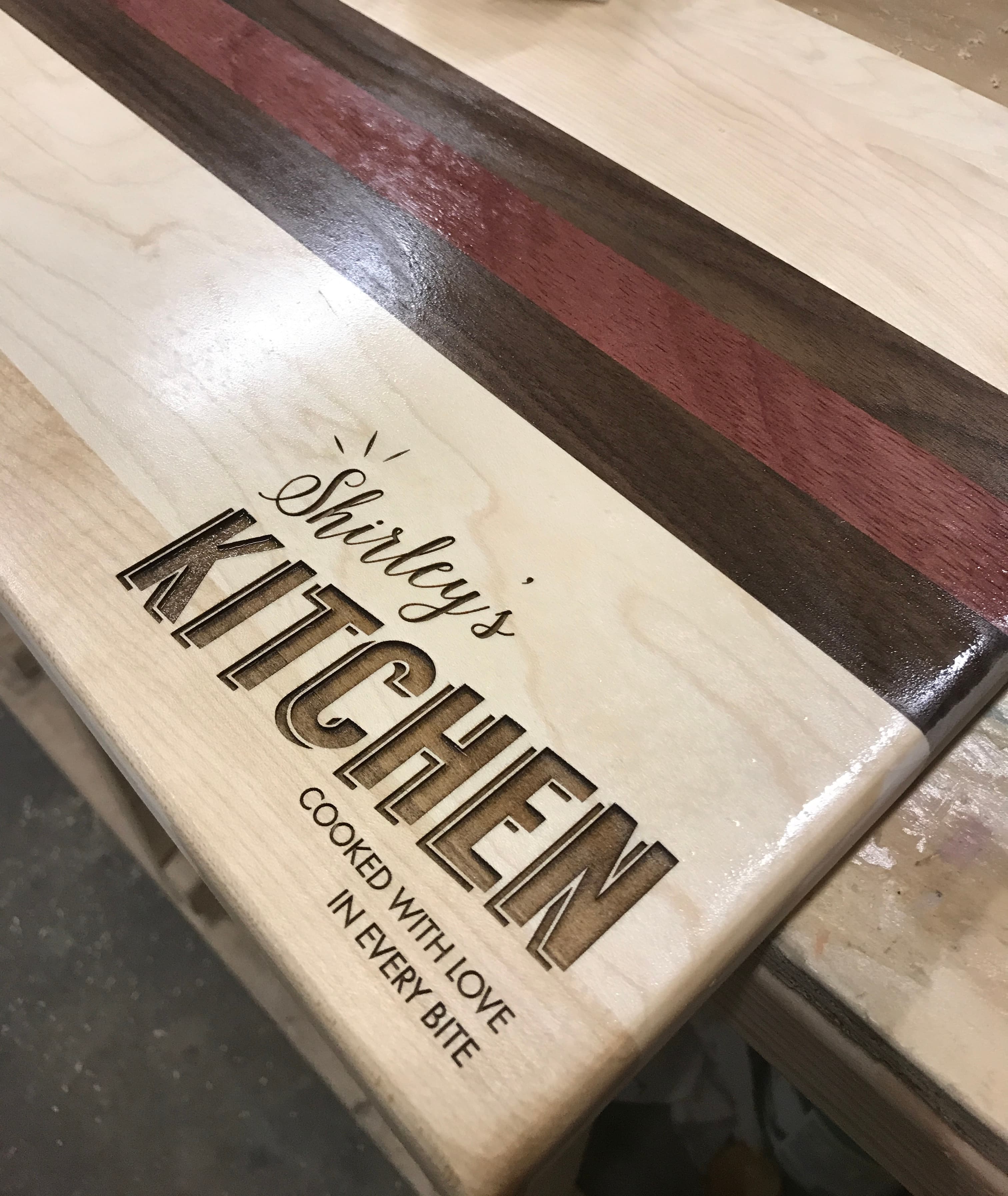 Finished custom engraved cutting board reading Shirley's Kitchen — Cooked With Love In Every Bite, featuring maple, walnut, and purple heart wood