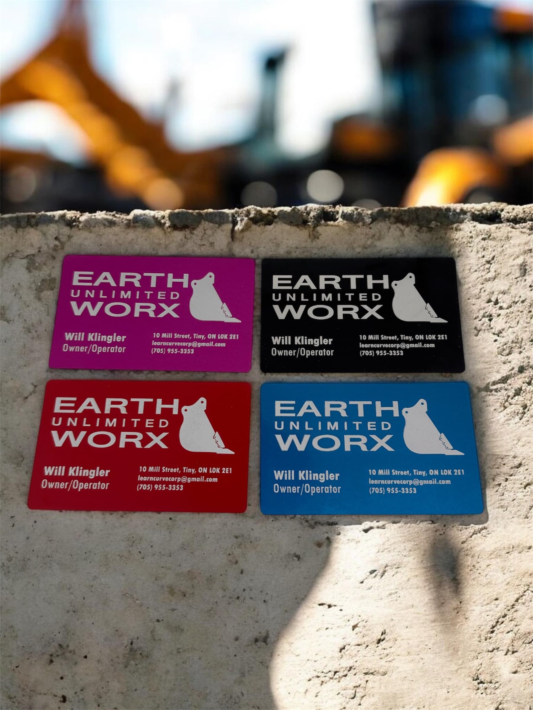 Four anodized aluminum business cards for Earth Unlimited Worx, in magenta, black, red, and blue, resting on a concrete ledge
